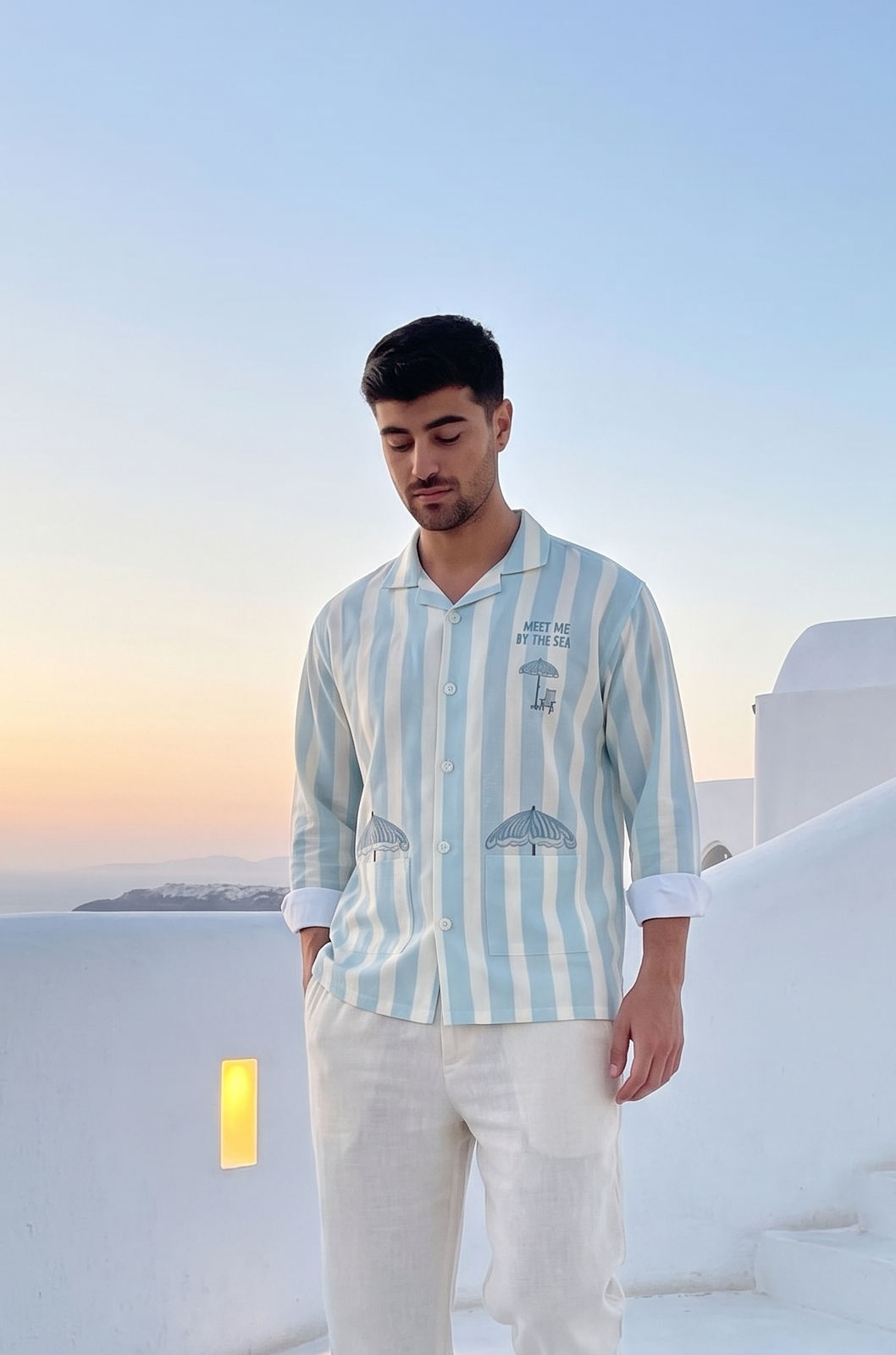 Man in a striped shirt and white pants standing on a white staircase with a blue sky and sunset in the background.
